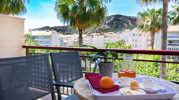 A balcony with a table set for breakfast, including a tray with orange juice, muffins, and a cup. There is a chair and a potted plant. - apartamentos Albir Confort (L'Alfas del Pi)