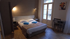 A neatly made bed with two pillows, a bedside table with a lamp, a chair, and a window with a view of the outside. - Le Céretan Hôtel (Ceret)