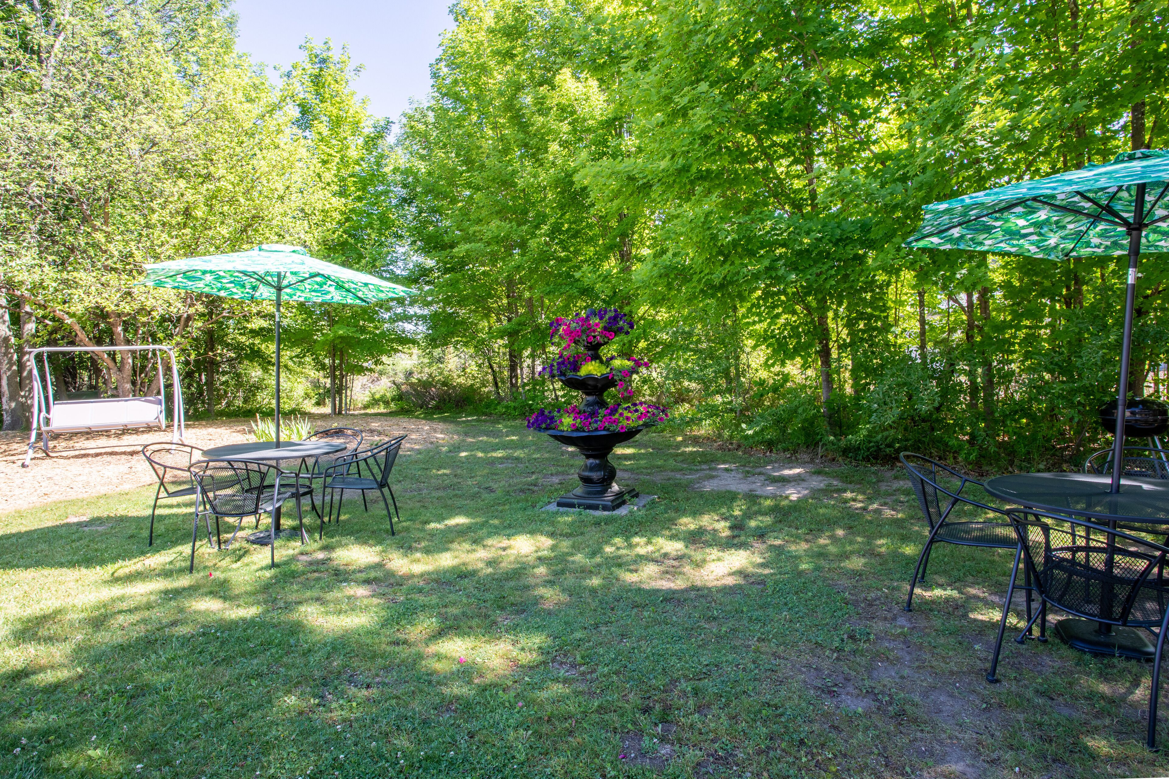 Un espace extérieur aménagé en terrasse, avec une table ronde et des chaises, entouré d’une végétation luxuriante et agrémenté de deux parasols verts qui offrent de l’ombre.