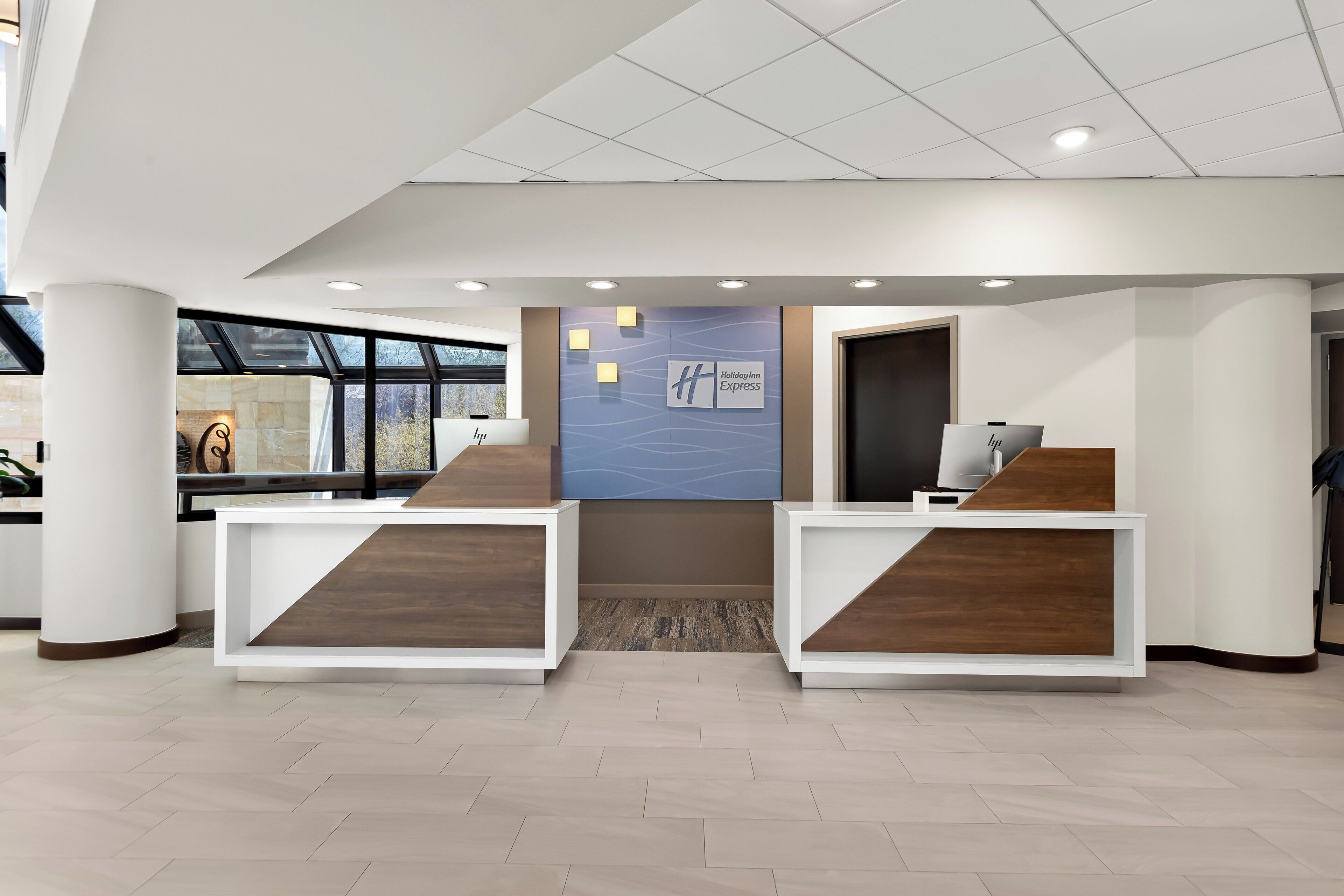 A modern hotel reception area with a large window, a reception desk, and a wall-mounted sign for Holiday Inn Express.