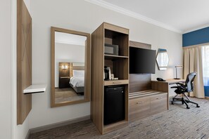 A hotel room with a wooden desk, a black office chair, a television mounted on the wall, and a small black refrigerator.