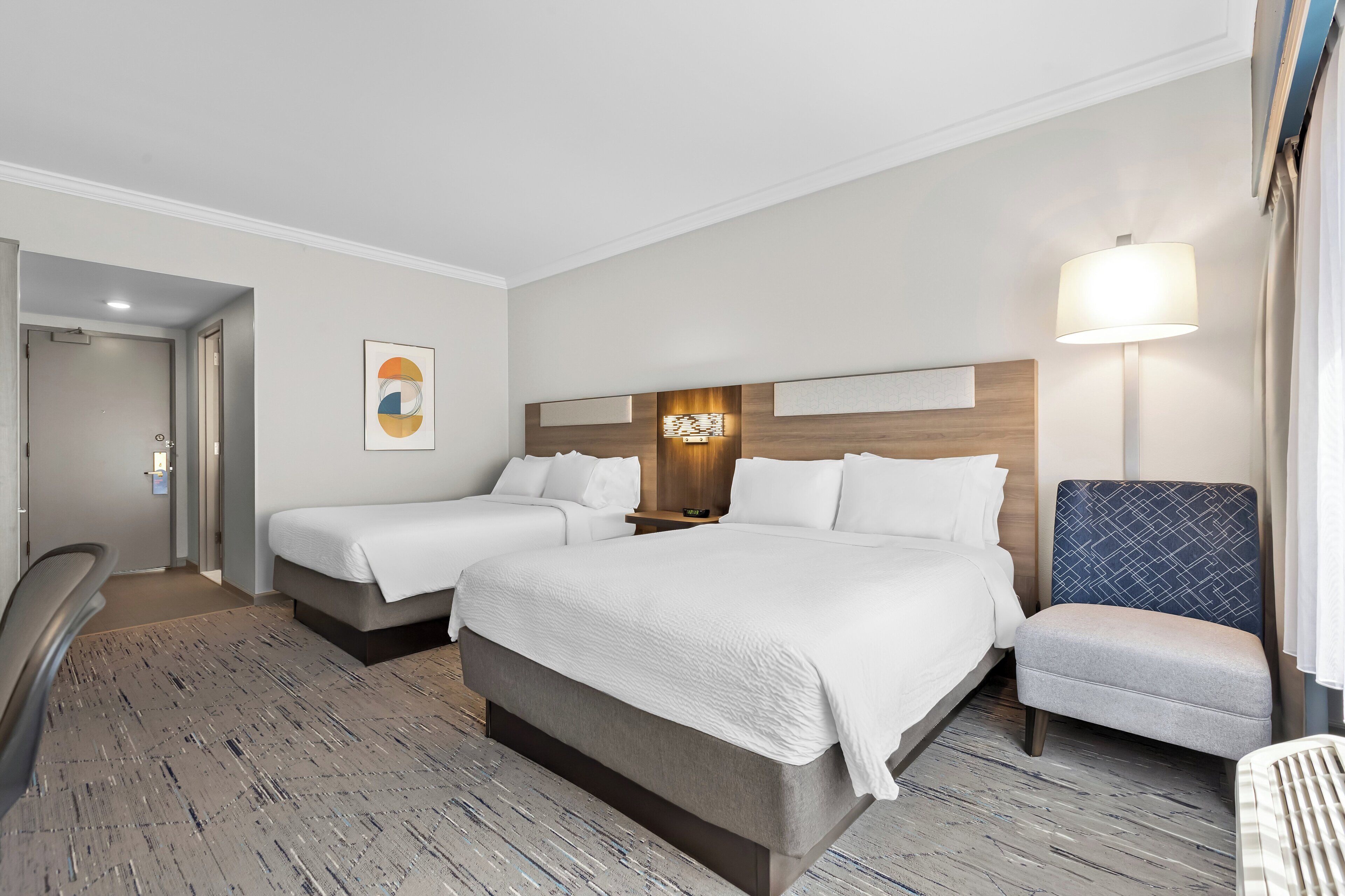 Une chambre d’hôtel avec deux lits, un bureau, une chaise et une table de chevet avec une lampe.