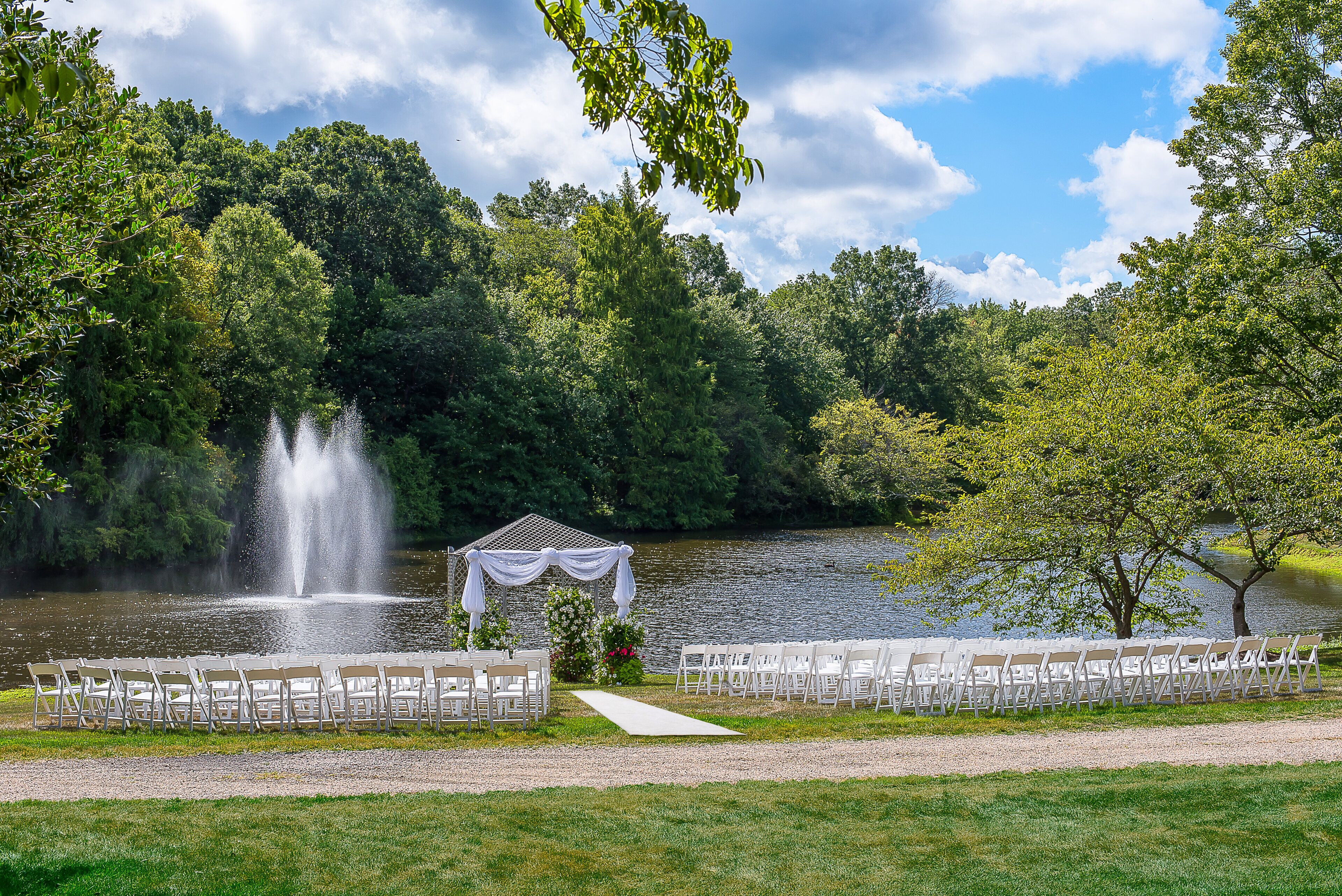 A gazebo with a water fountain, surrounded by a gravel path and white chairs, set against a backdrop of trees and a lake.