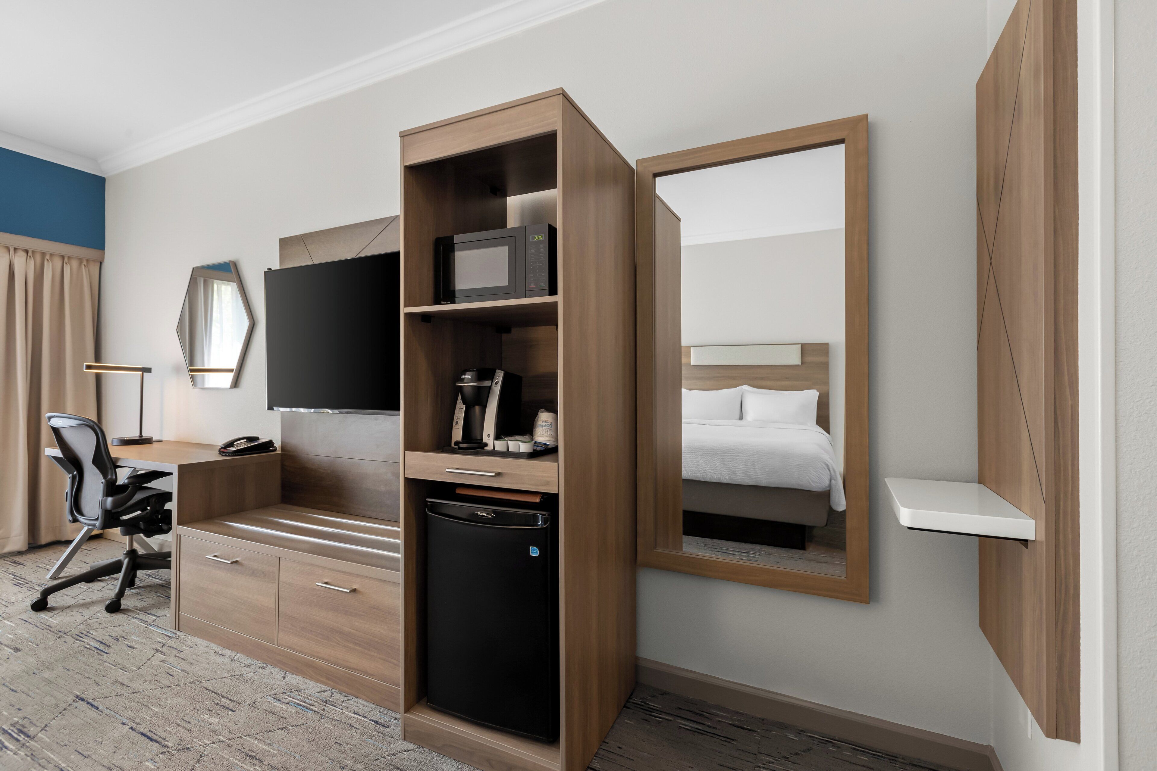 Hotelkamer met een houten bureau, een zwarte koelkast, een flatscreen televisie, een bed met witte lakens en een grote spiegel.