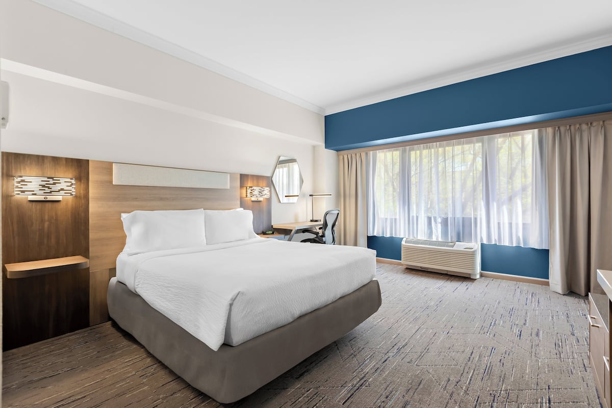 A modern hotel room with a large bed, a desk, a chair, and a window with curtains.