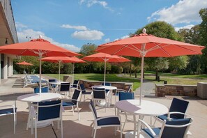 An outdoor patio area with round tables and chairs, red umbrellas, and a view of a golf course.