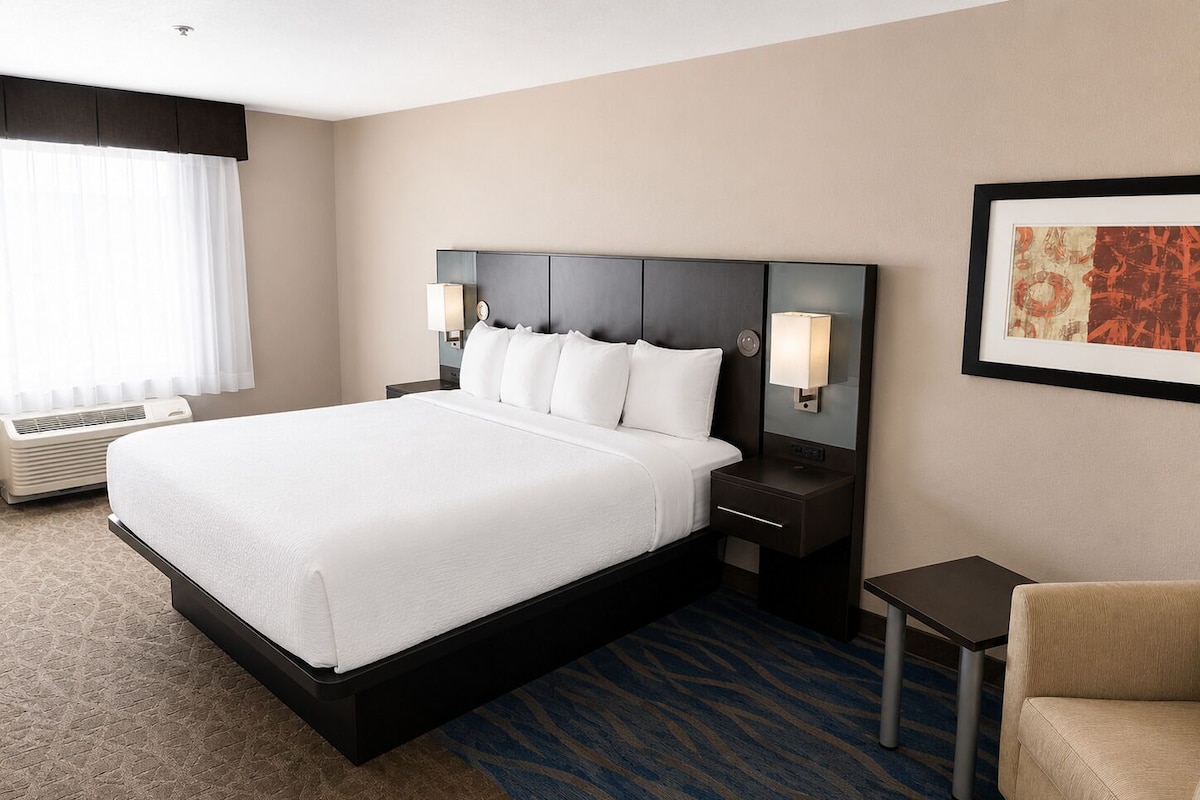 A hotel room with a large bed, two bedside tables, a chair, a wall-mounted lamp, and a framed picture.