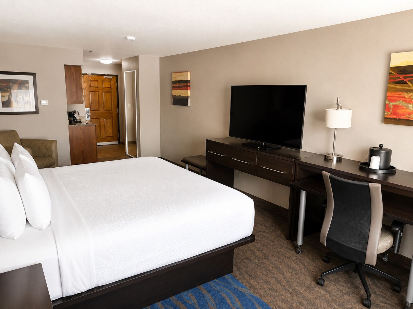 A hotel room with a bed, a desk with a flat-screen TV, a chair, a lamp, and a painting on the wall.