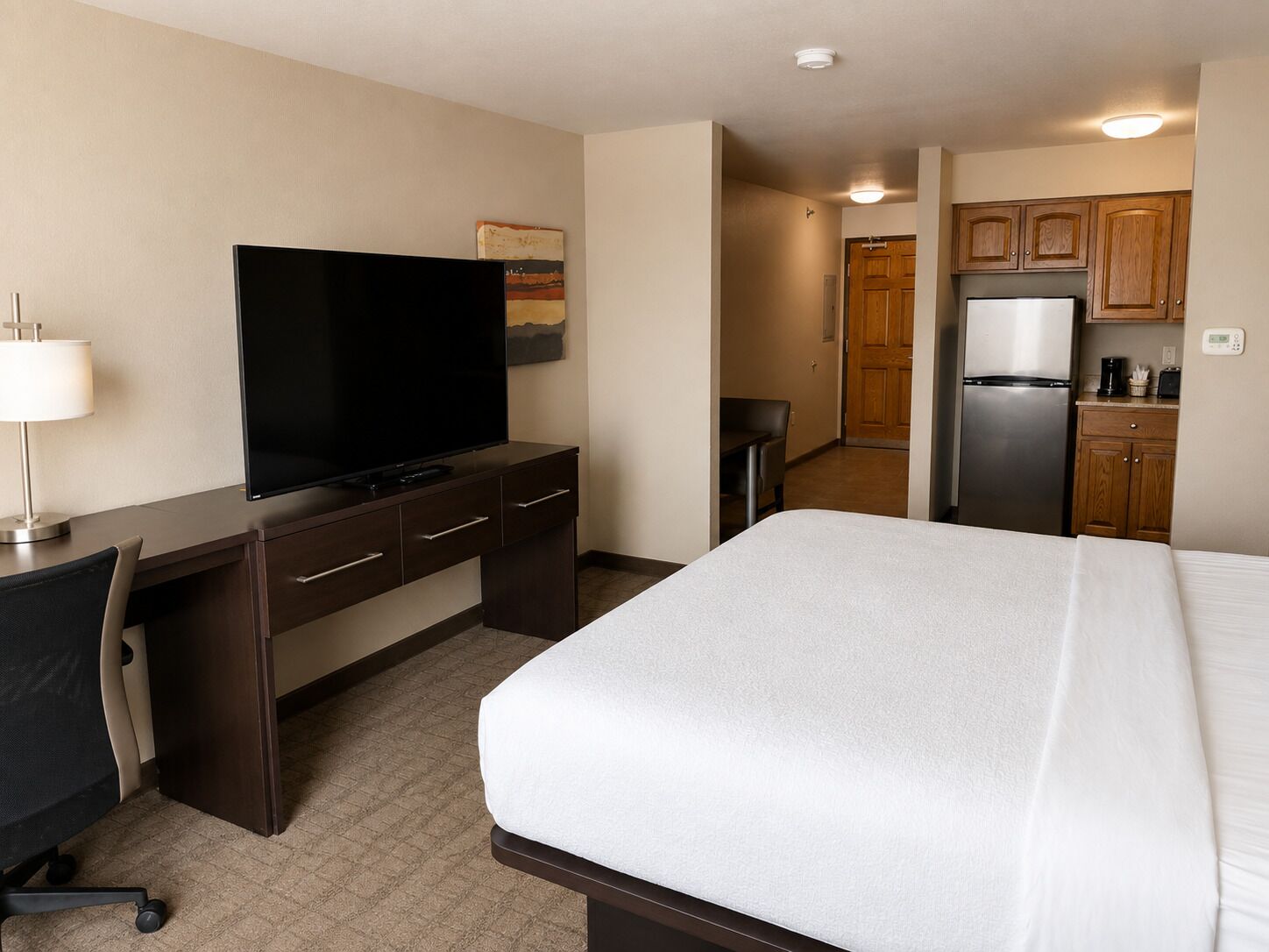 A hotel room with a bed, a desk with a television, a chair, a small table, and a kitchenette area.