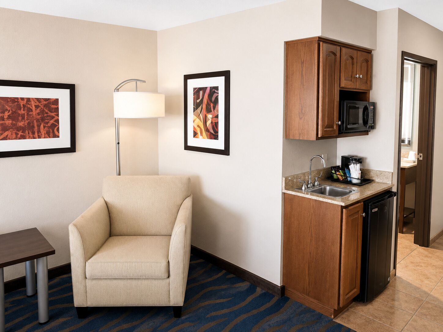 A hotel room with a kitchenette, a chair, a lamp, and two framed pictures on the wall.