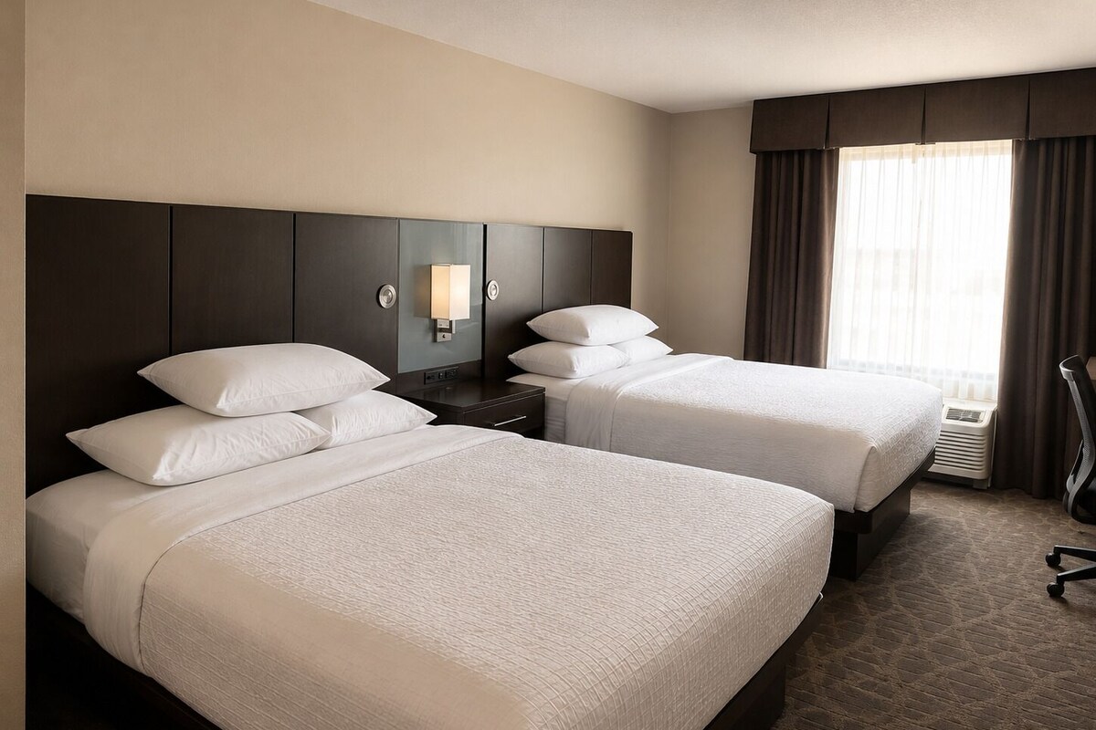 A hotel room with two beds, a nightstand, a lamp, a chair, and a window with curtains.