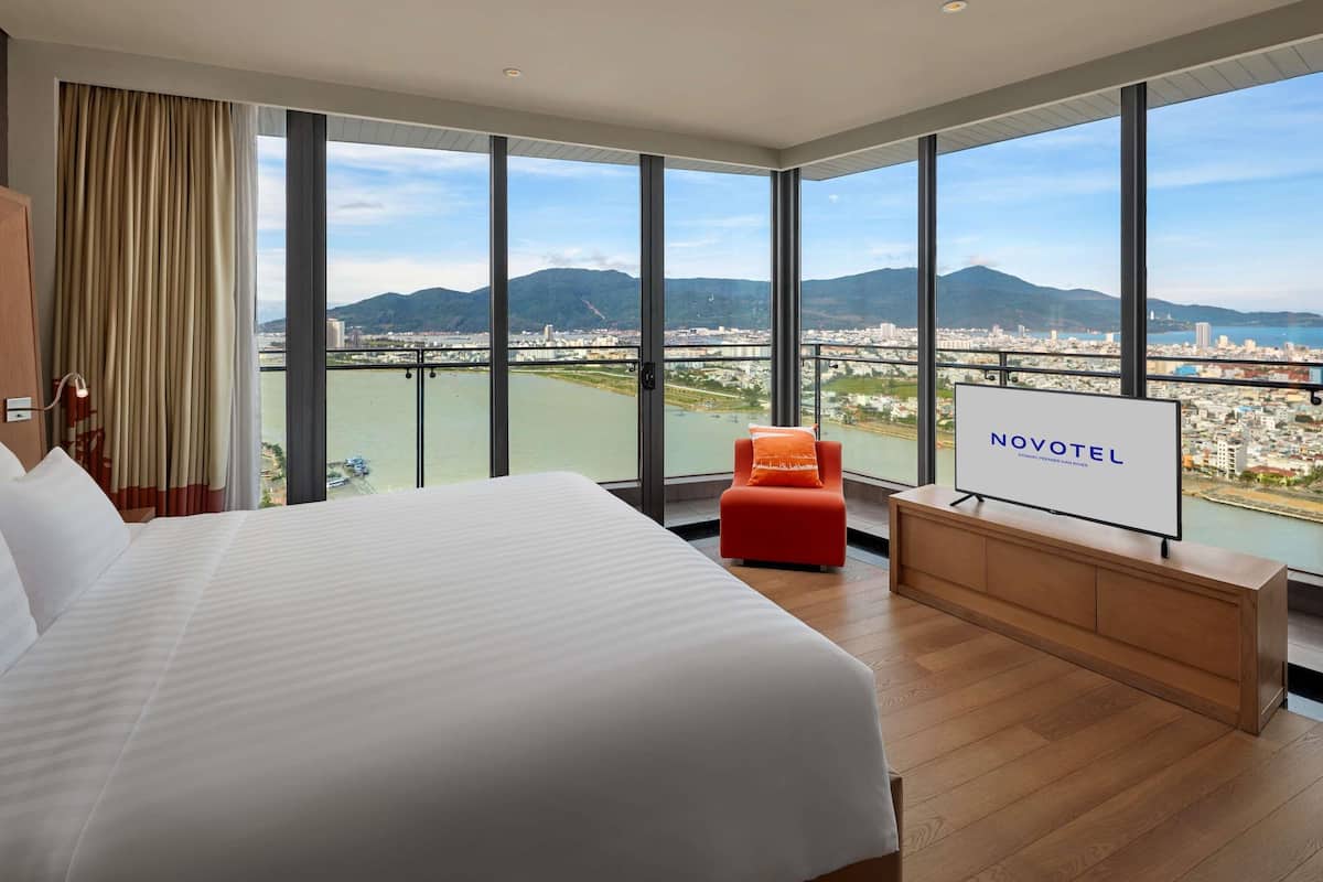 Habitación de hotel con una cama grande, televisión, escritorio de madera y vistas a la ciudad y las montañas.