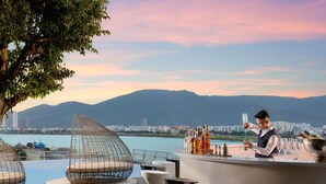 Uma área de bar com um bartender, uma área de estar à beira da piscina com cadeiras modernas e uma vista panorâmica de montanhas e um corpo d'água.