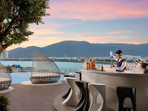 Zona de bar con un barman, área de estar junto a la piscina con sillas modernas y una vista panorámica de montañas y una masa de agua.