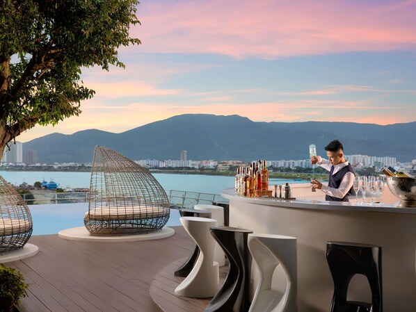 Zona de bar con un barman, área de estar junto a la piscina con sillas modernas y una vista panorámica de montañas y una masa de agua.