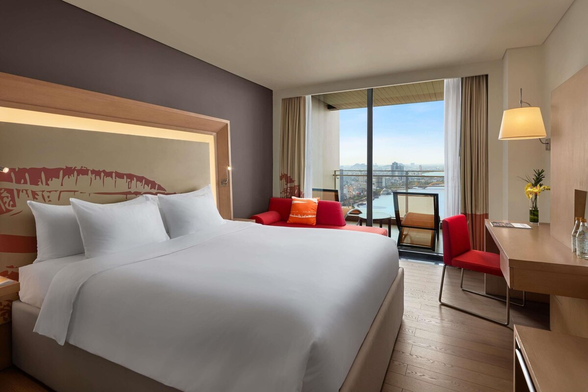 A hotel room with a large bed, a desk, a chair, and a view of a cityscape through large windows.