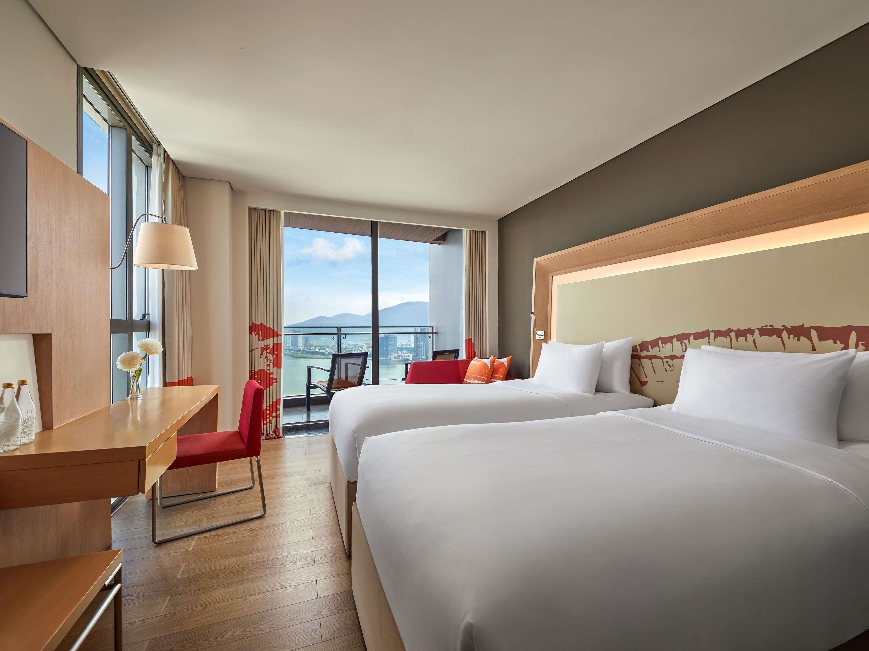 A hotel room with two beds, a desk, a chair, and a view of the city.