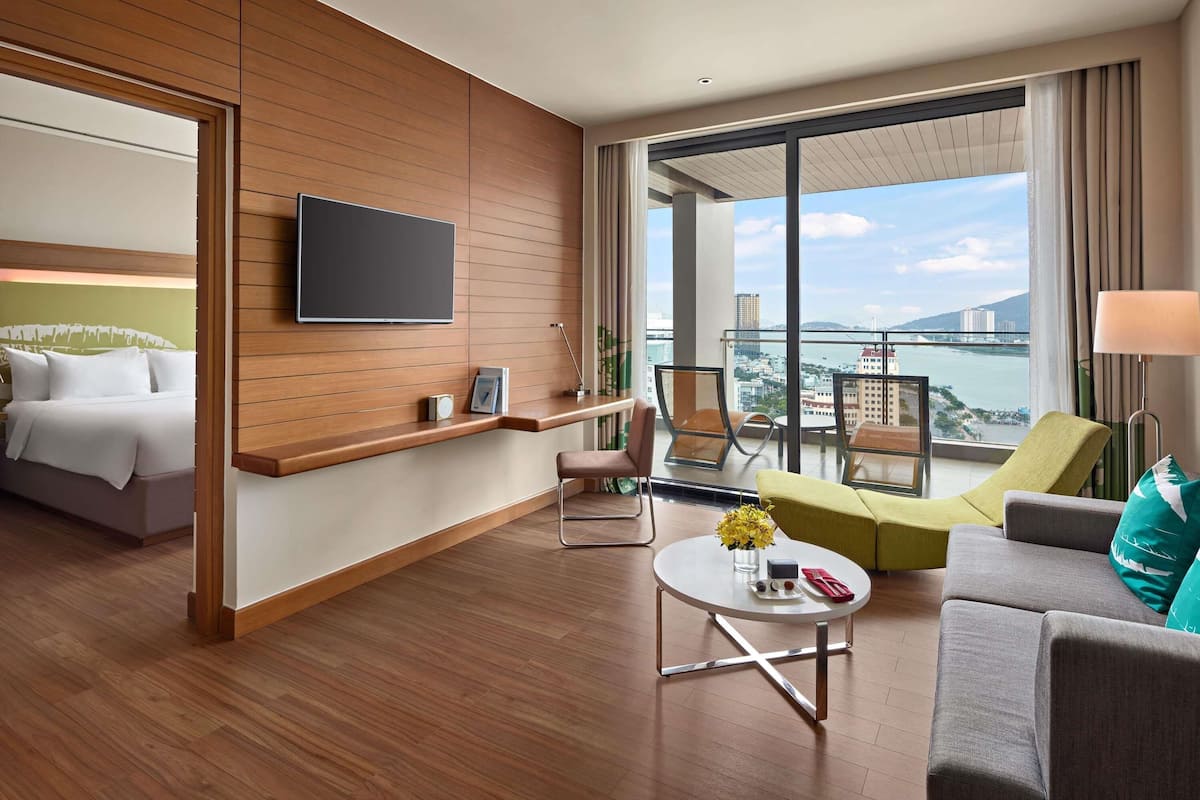 Habitación de hotel con cama, televisión, balcón con vistas, zona de estar con mesa y sofá.