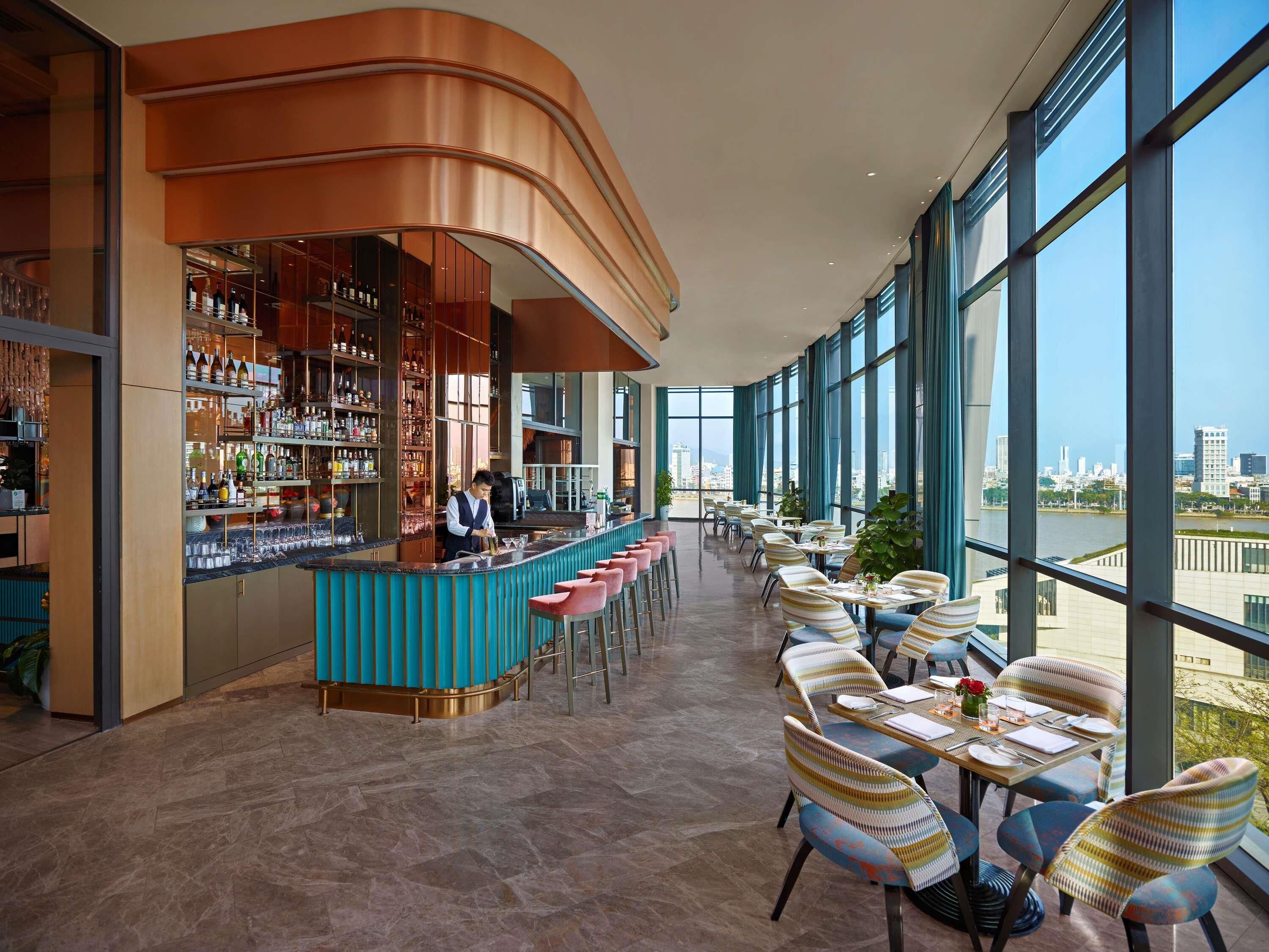 A modern bar area with a curved counter, a bartender, and a view of the city through large windows.