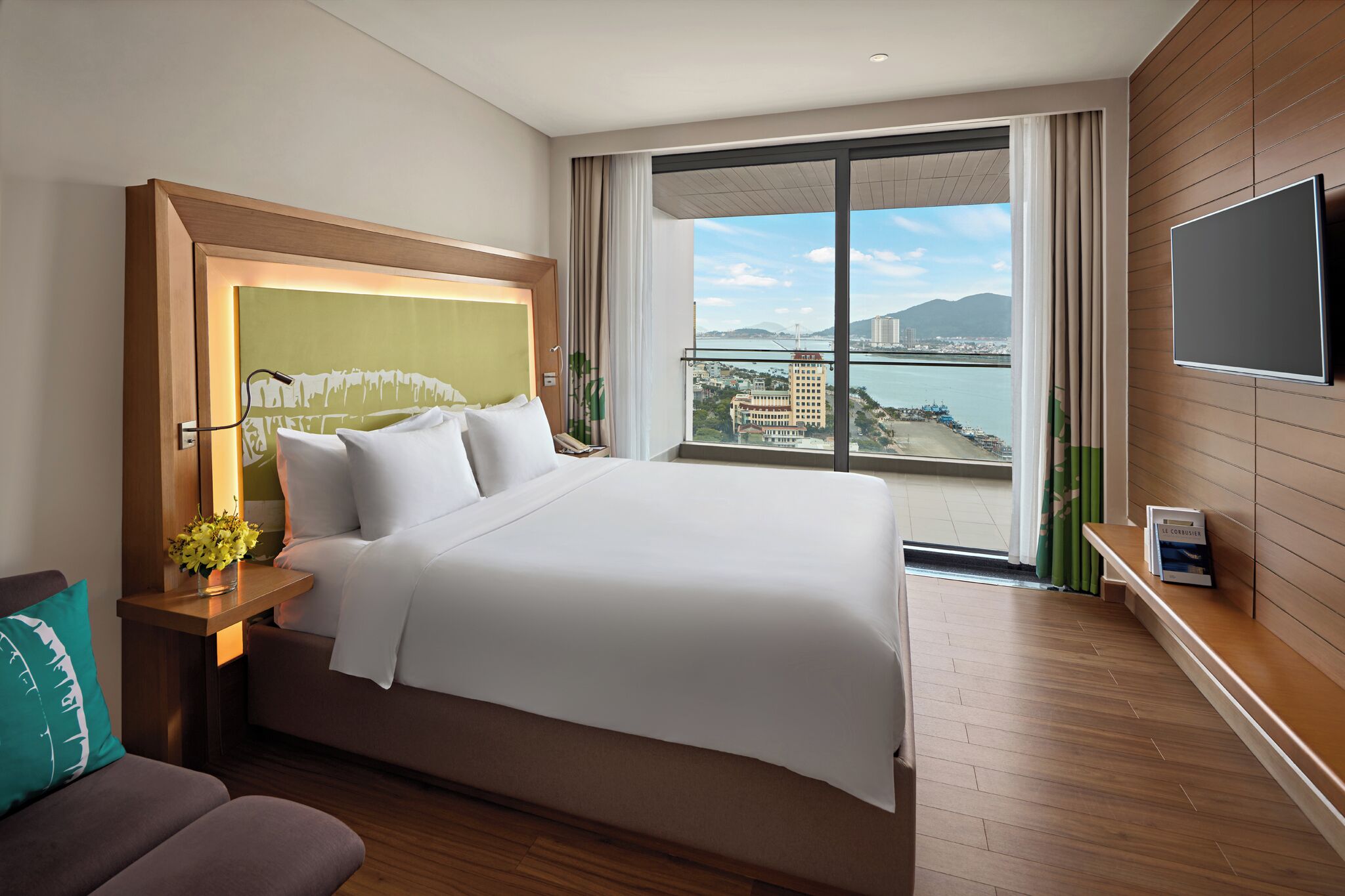 A hotel room with a large bed, a TV, a balcony with a city view, and a wooden floor.