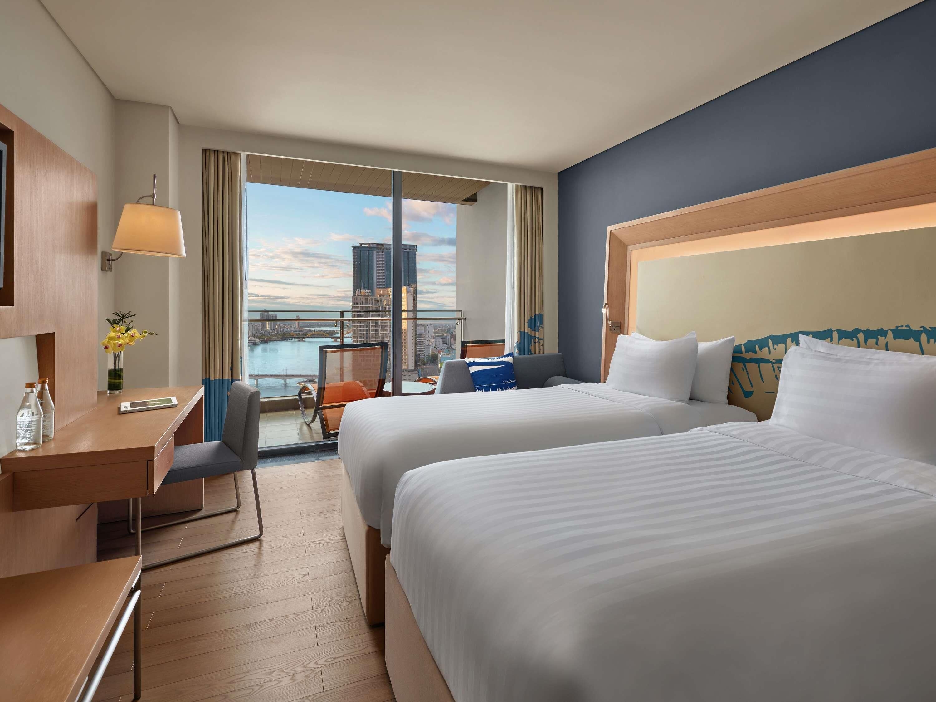 A hotel room with two beds, a desk, a chair, a small table, and a view of a cityscape.