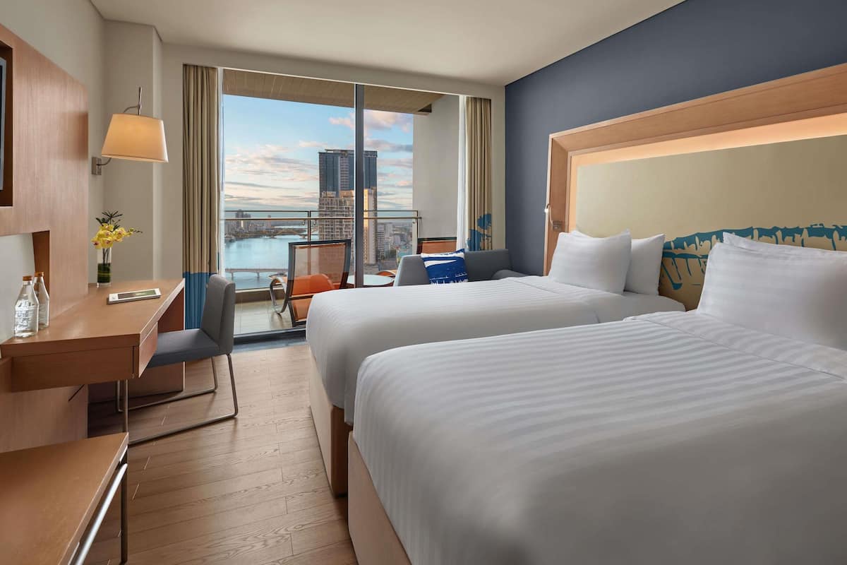A hotel room with two beds, a desk, a chair, a small table, and a view of a cityscape.