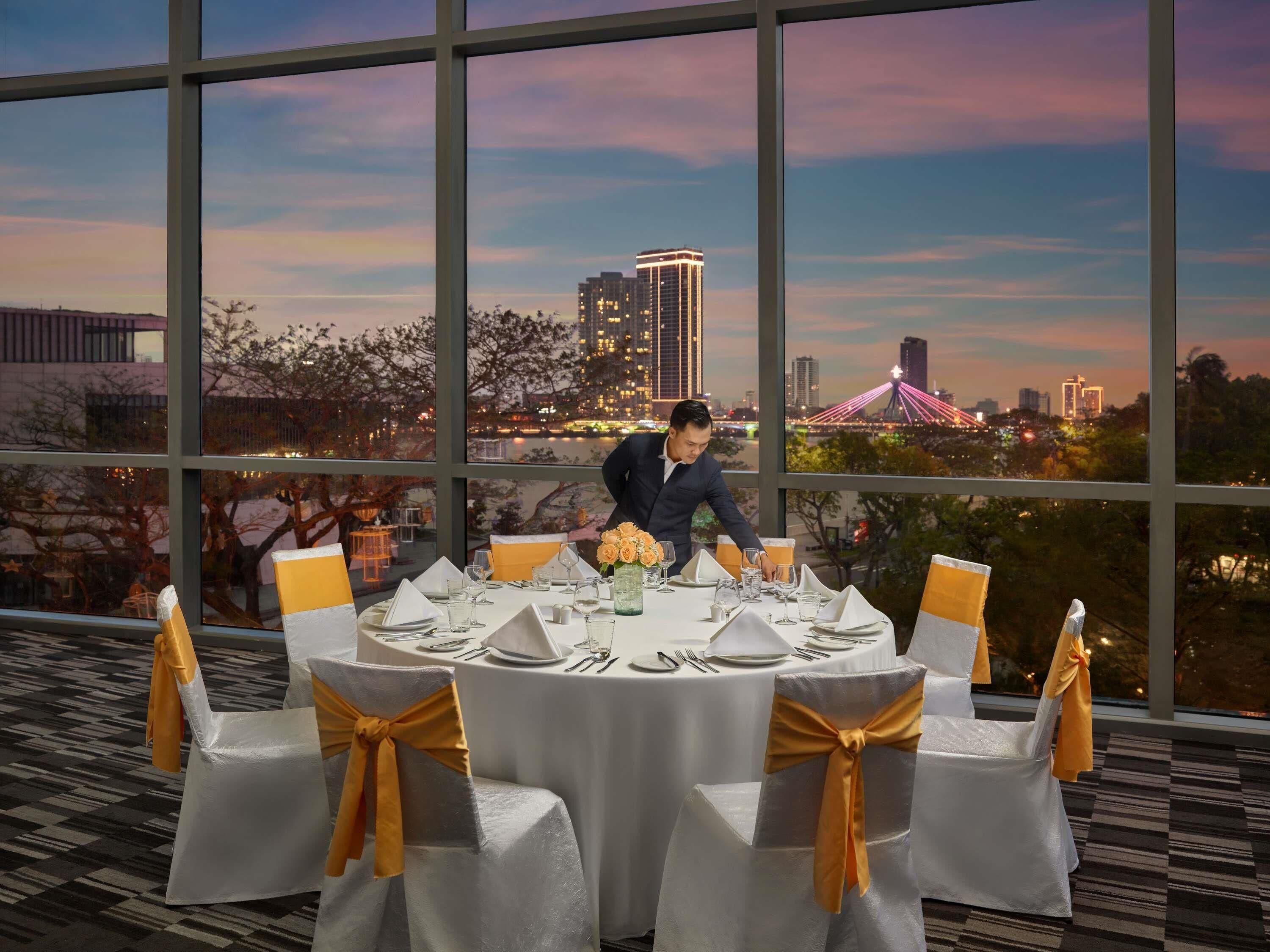A dining area with round tables set for a meal, large windows with a cityscape view, and a person arranging the table settings.