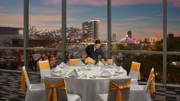 A dining area with round tables set for a meal, large windows with a cityscape view, and a person arranging the table settings.