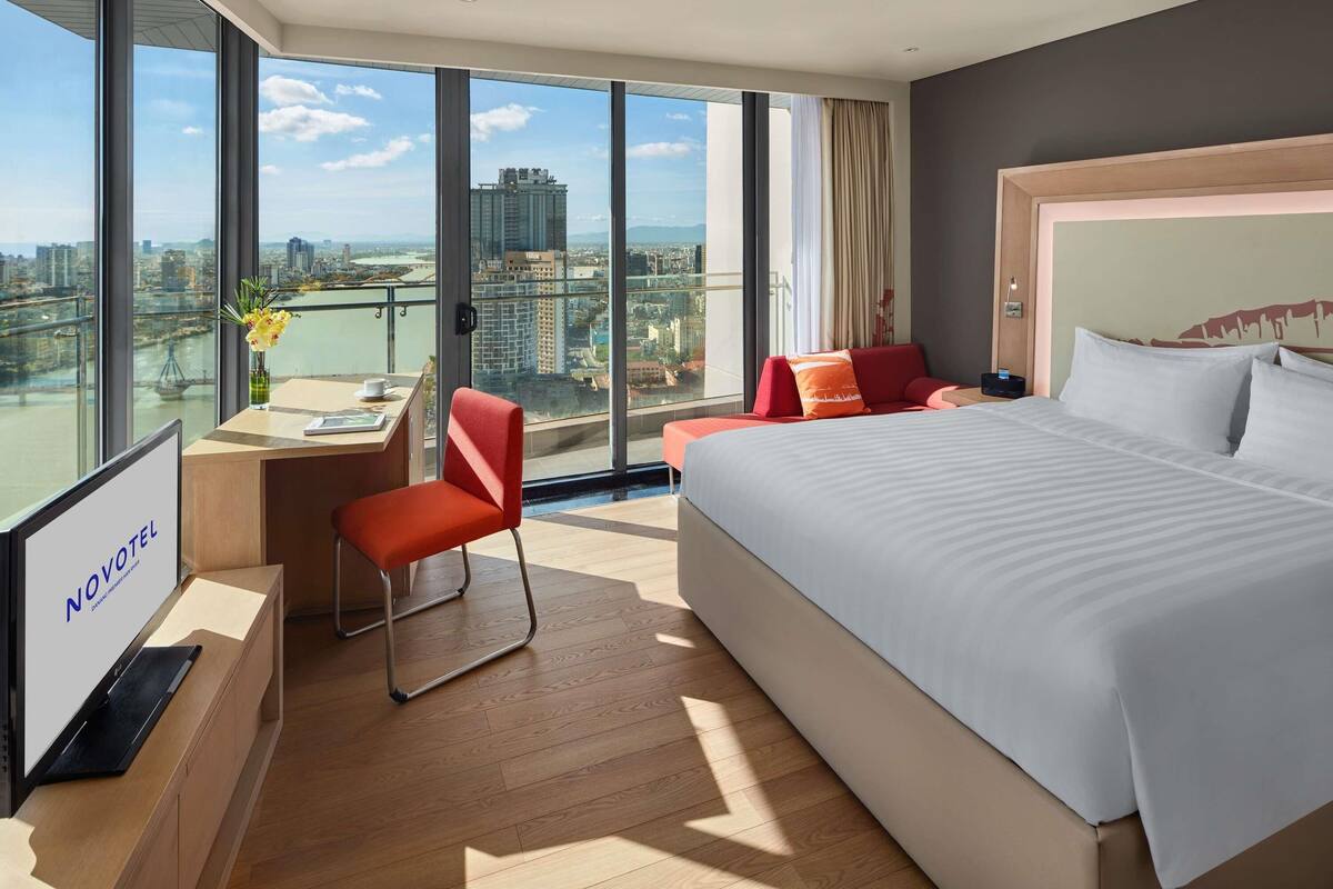 Una habitación de hotel moderna con una cama grande, un escritorio con una silla y vistas a la ciudad a través de amplias ventanas.