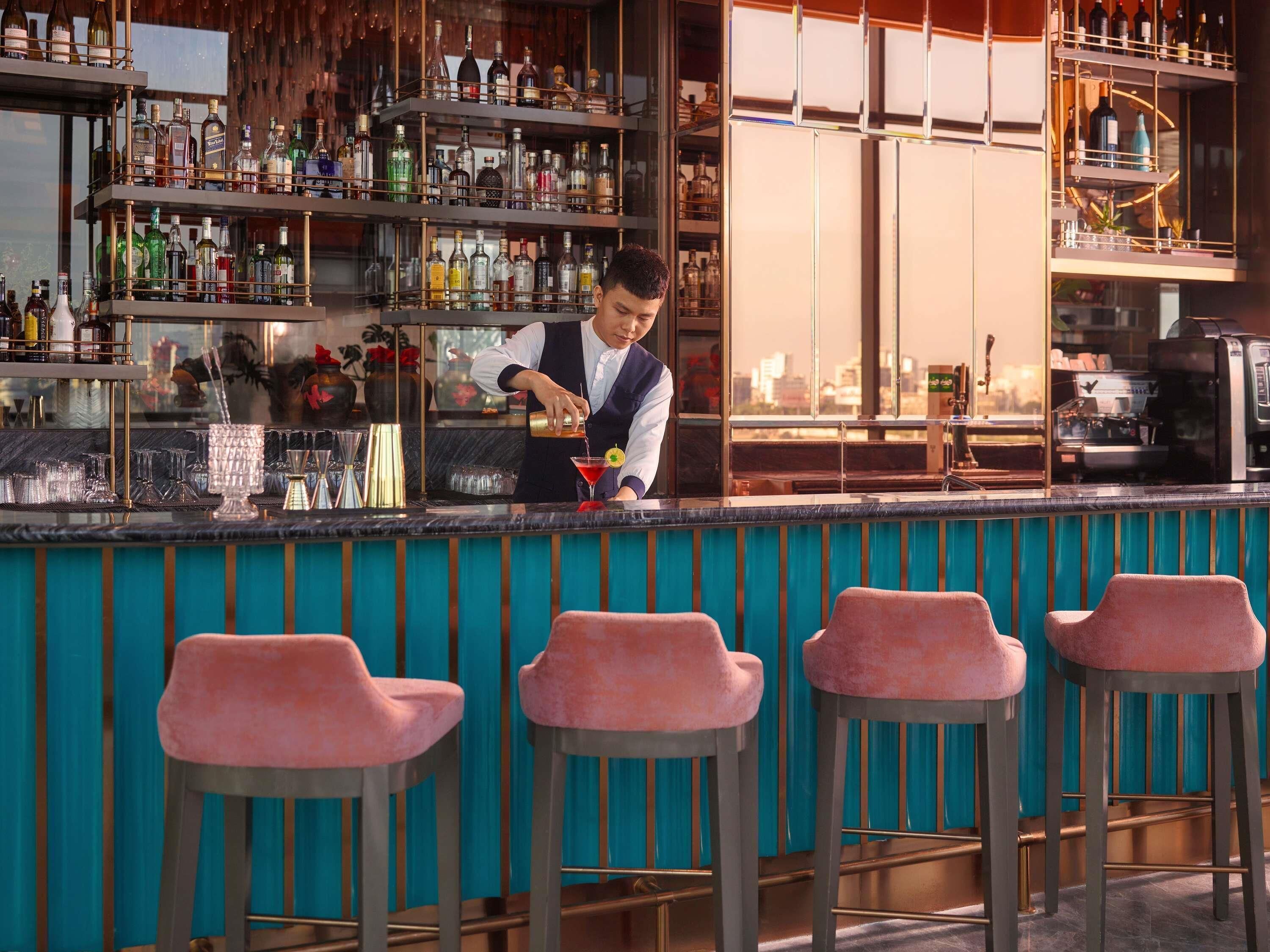 A bar area with a bartender preparing a drink, shelves with bottles, and bar stools.