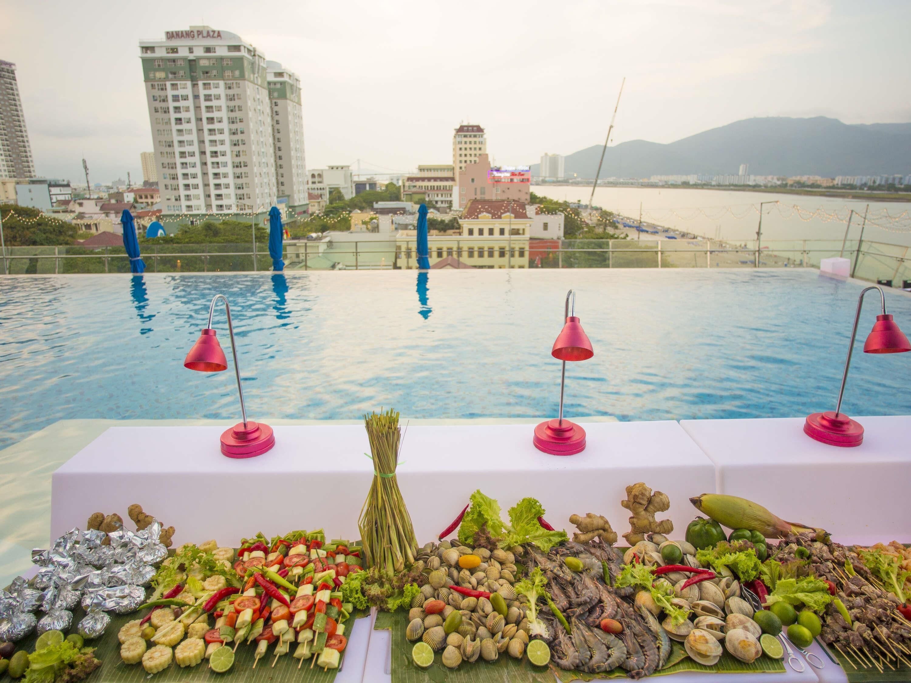 A poolside table with food and drinks, overlooking a cityscape with buildings and a body of water.