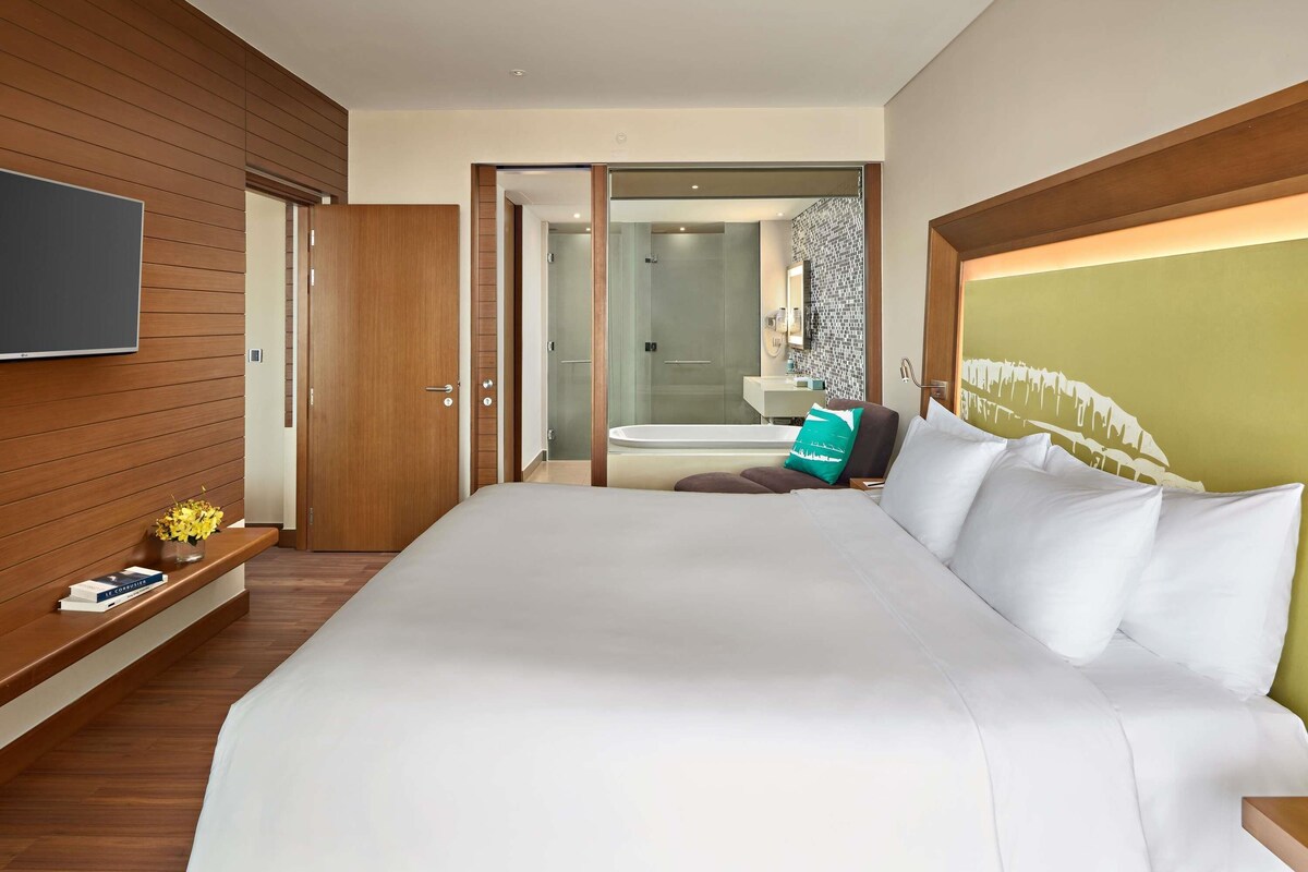A modern hotel room with a large bed, wooden panel walls, a flat-screen TV, and a bathroom with a glass shower.