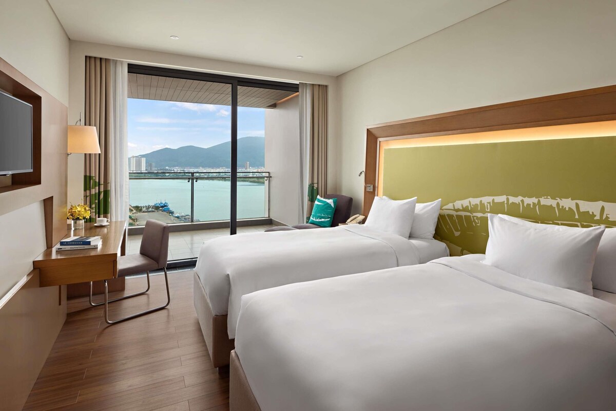 A hotel room with two beds, a desk with a lamp, a chair, and a view of a body of water and mountains.