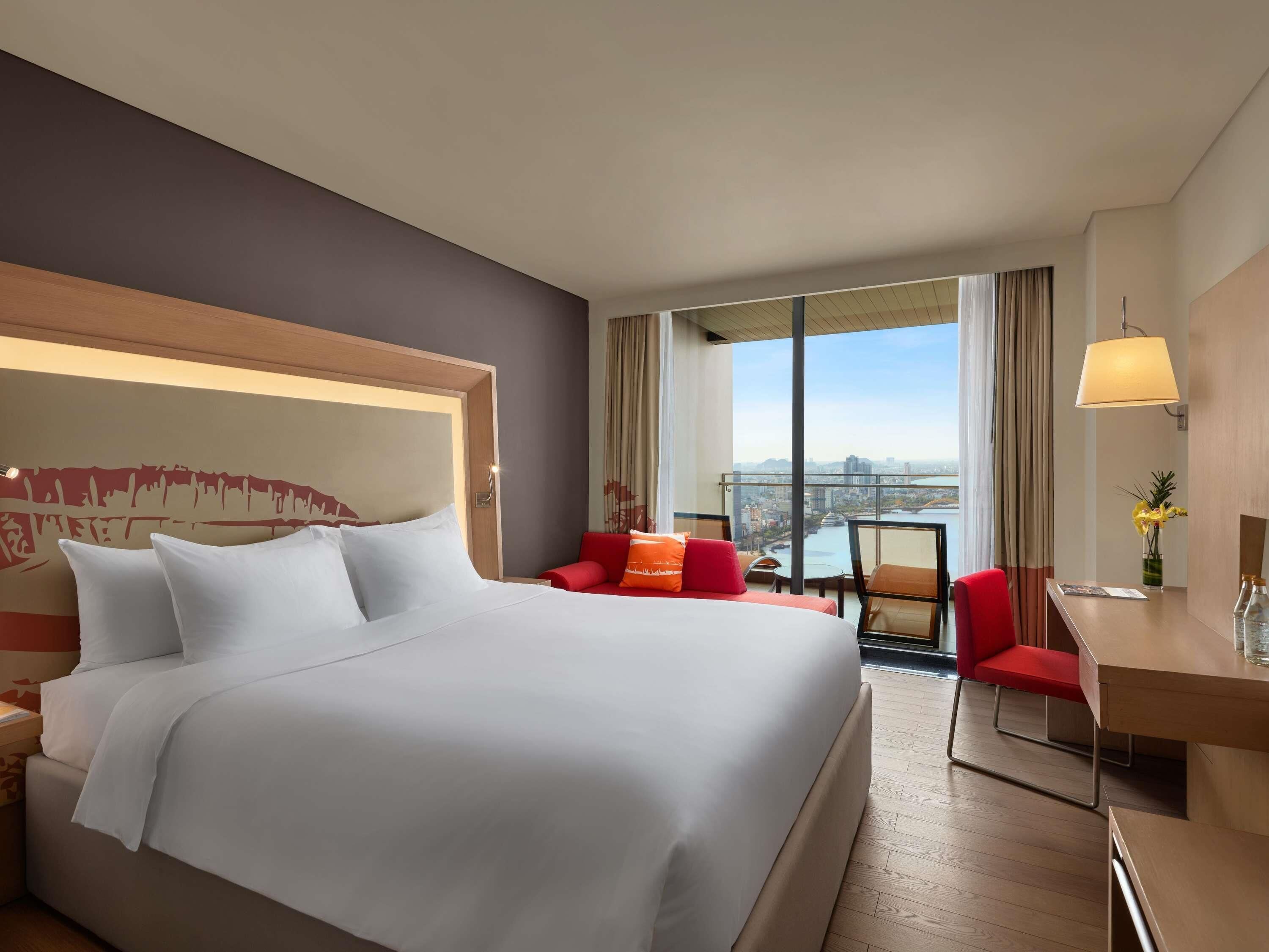 A hotel room with a large bed, a desk, a chair, and a view of a cityscape through large windows.