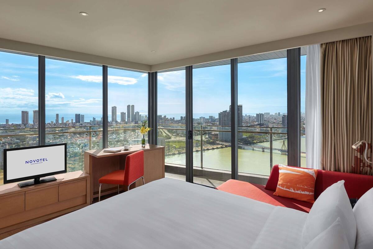 A hotel room with a large bed, a desk with a computer, a chair, and a TV displaying 'NOVOTEL'.