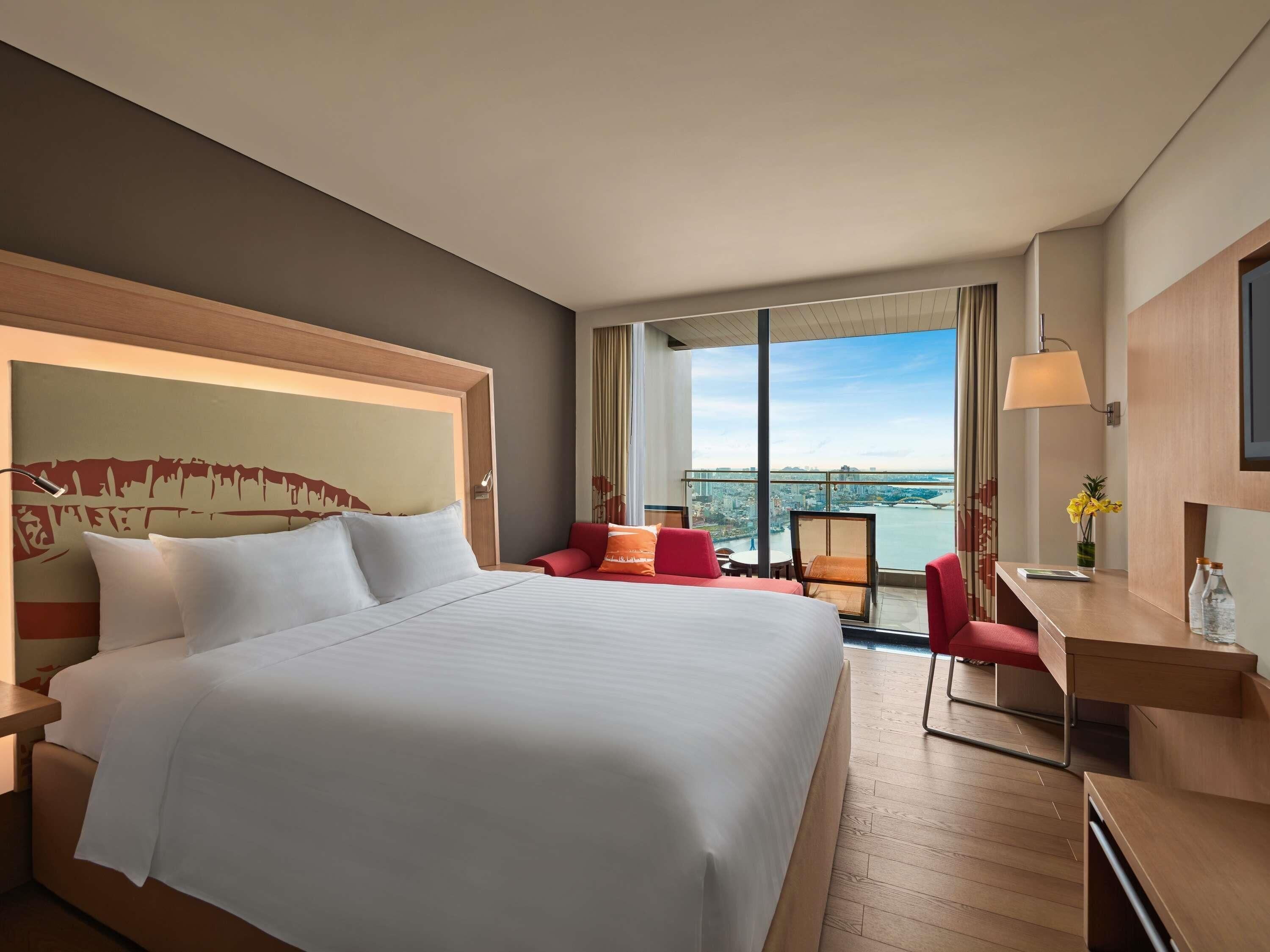 Habitación de hotel con una cama grande, un escritorio, una silla y vistas a un paisaje urbano.