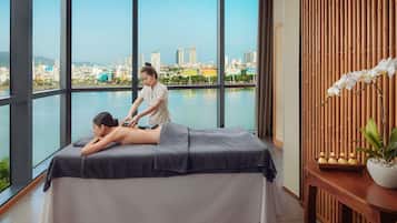 A massage therapy room with a city view, a massage table, and a wooden side table with a potted orchid.