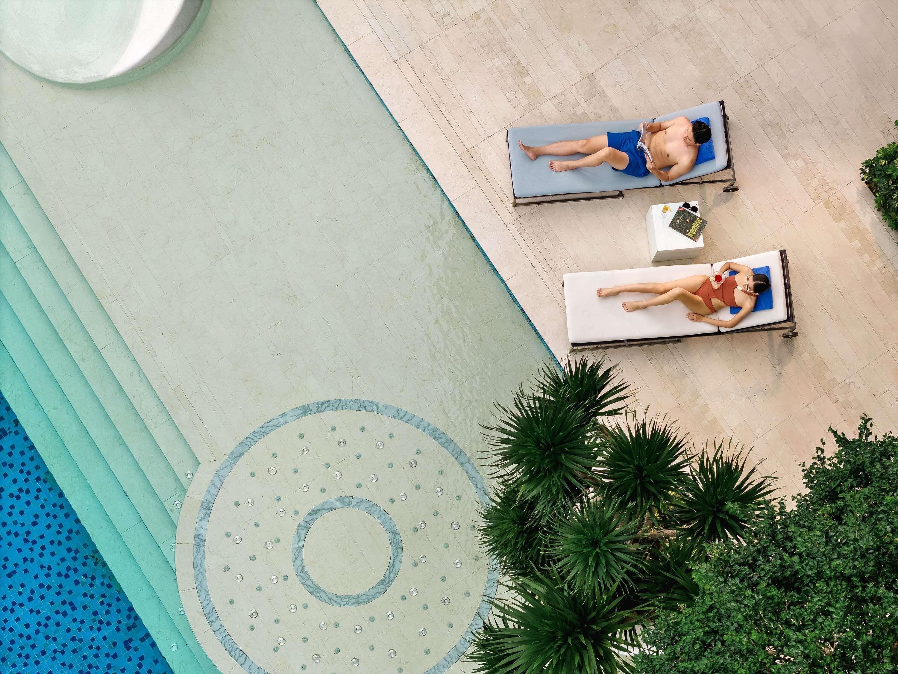 Two sun loungers by a poolside with a circular water feature and palm trees.