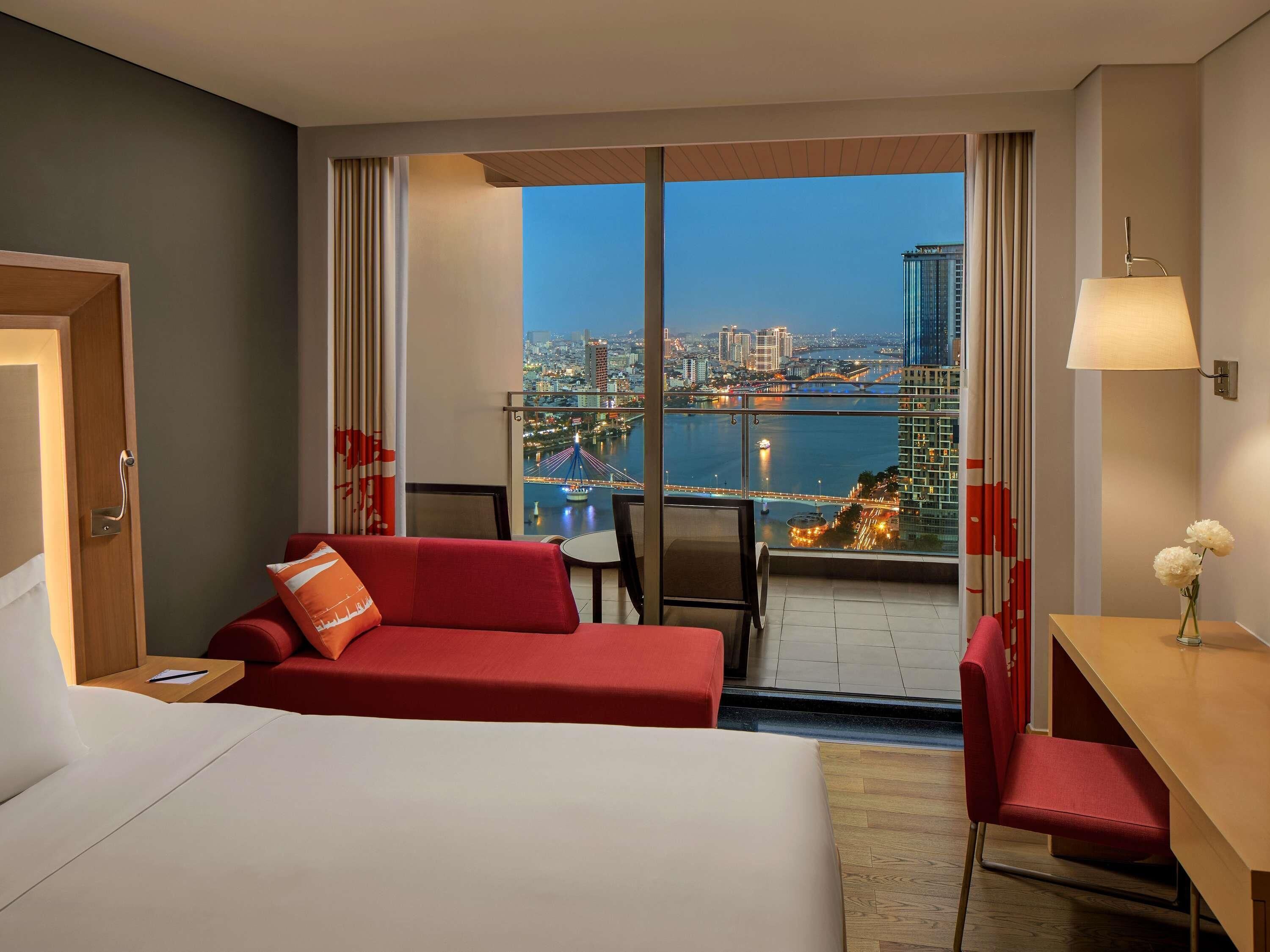 A hotel room with a bed, a red sofa, a desk, and a view of a cityscape and river.