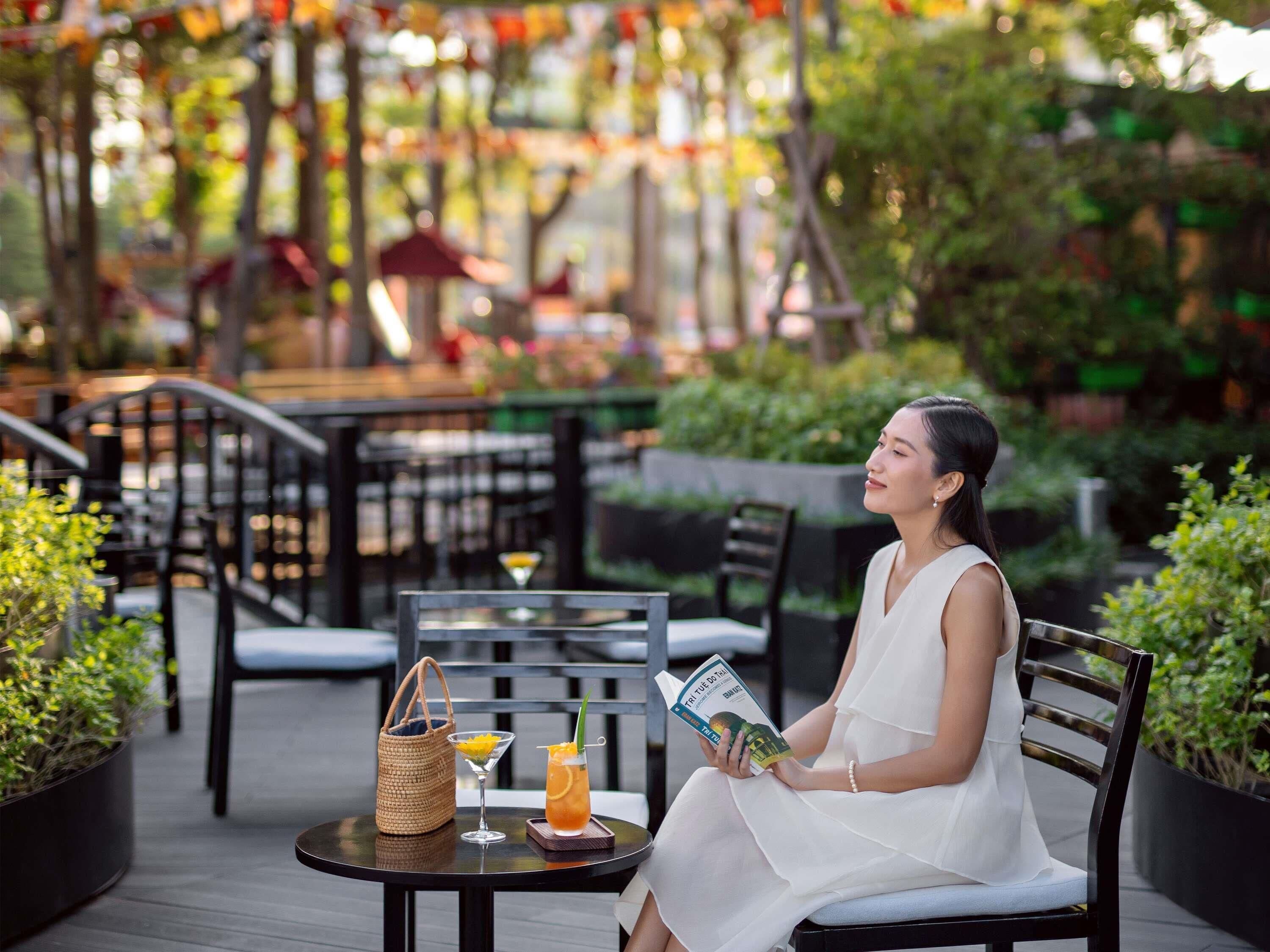 A woman seated at an outdoor table with a book, a drink, and a bag.