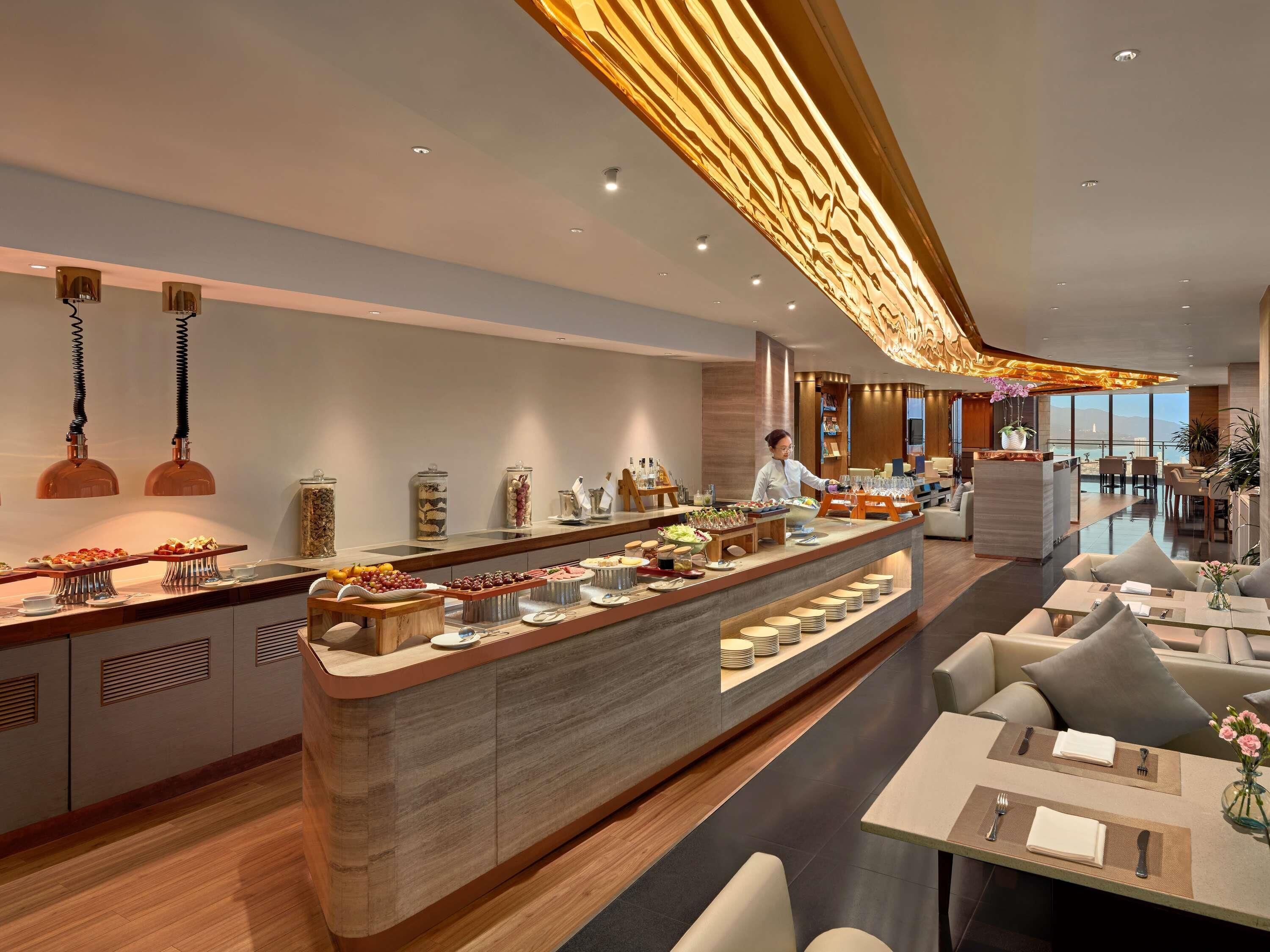 A modern hotel lobby with a central buffet, seating area, and a view of the sea.