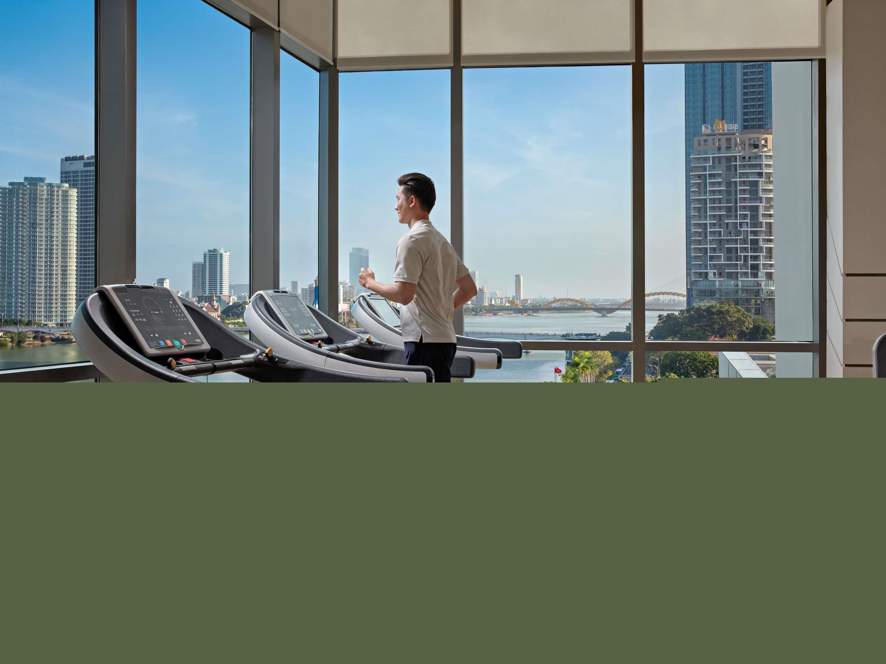 A man using a treadmill in a room with large windows overlooking a cityscape.