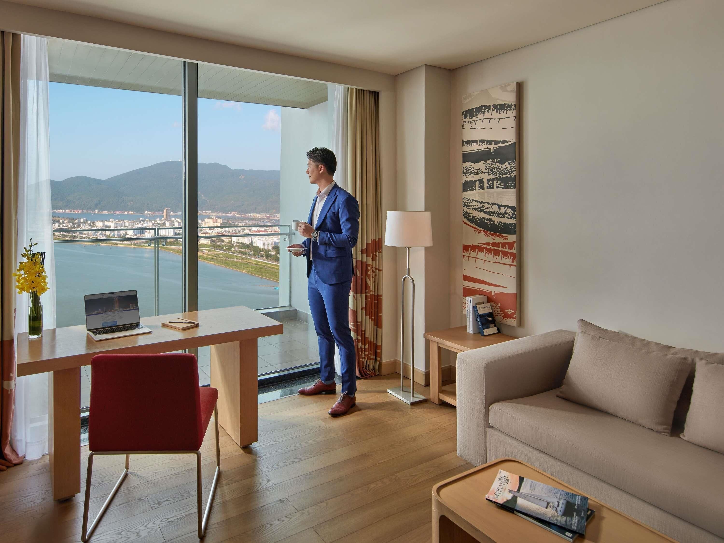 A hotel room with a large window, a desk with a laptop, a red chair, a sofa, and a lamp.