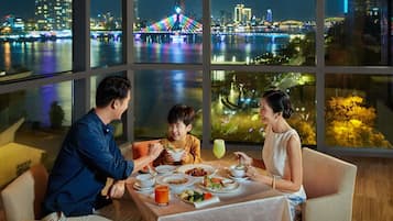 A dining room with a table set for three, large windows with a city view, and a table with various dishes and drinks.
