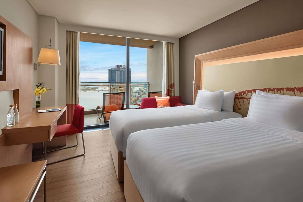 Habitación de hotel con dos camas, un escritorio, una silla y vistas a un paisaje urbano.