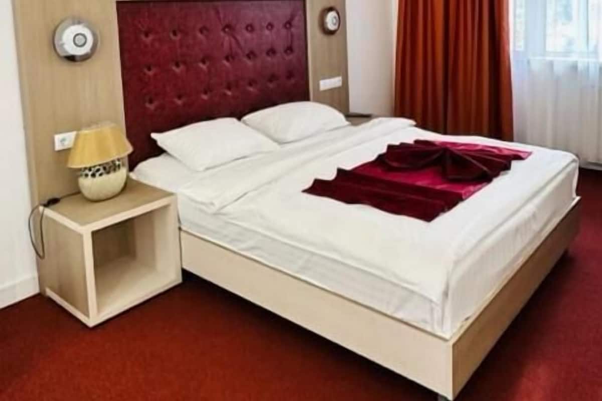 A hotel room with a large bed, a red headboard, a bedside lamp, and a window with curtains.