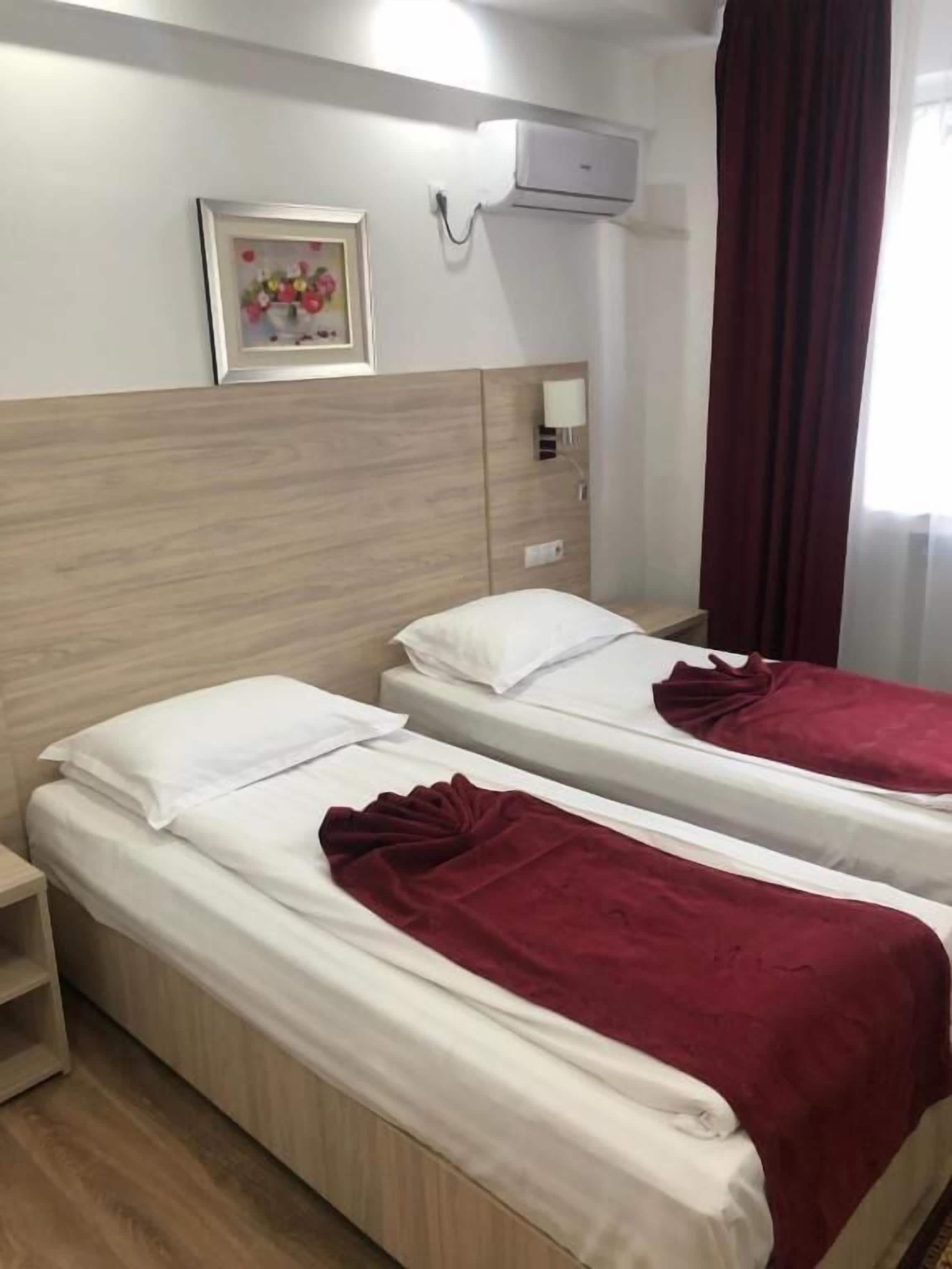 A hotel room with two beds, each with white linens and a red blanket. There is a framed picture on the wall, a bedside table with a lamp, and an air conditioning unit above the beds.