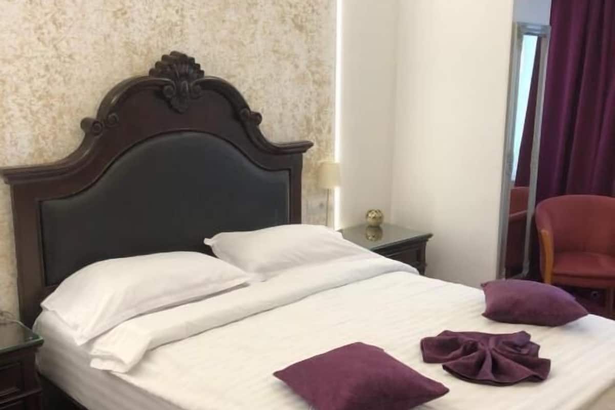 A neatly made bed with white linens and purple pillows, a wooden headboard, a nightstand with a lamp, and a red chair in a room with textured walls and a window with purple curtains.