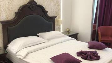 Una cama bien hecha con sábanas blancas y almohadones morados, un cabecero de madera, una mesita de noche con una lámpara y una silla roja en una habitación con paredes con textura y un ventanal con cortinas moradas.