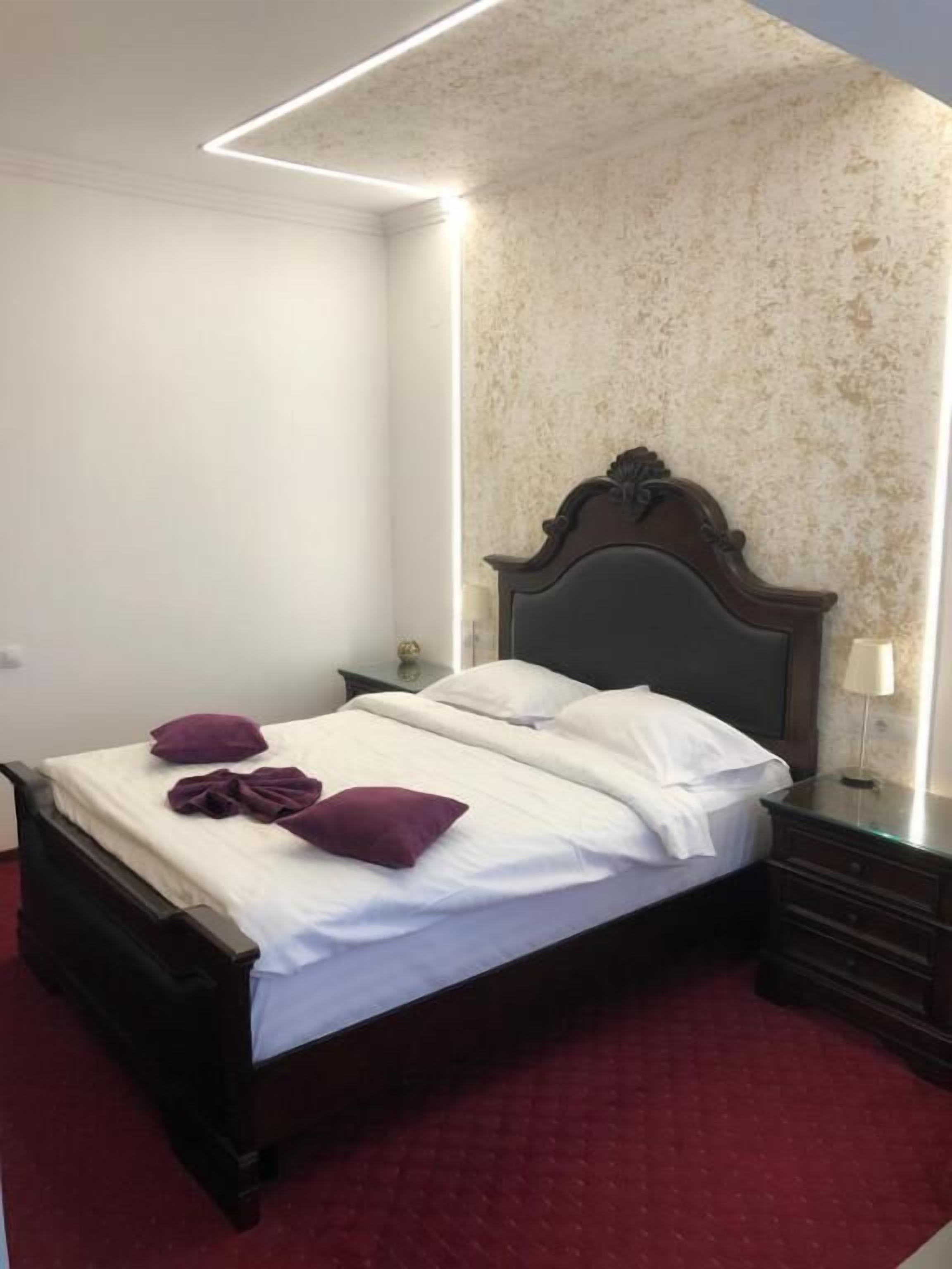 A neatly made bed with white linens and purple pillows, a dark wooden headboard, and a red carpeted floor.